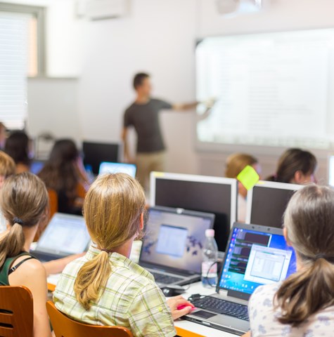 An image of a man pointing to a whiteboard giving a presentation to students sat at their desks